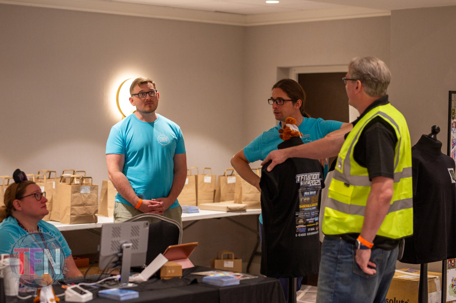 Several TFNation organisers and volunteers, talking at the merch desk. One is wearing a Hi Vis safety vest